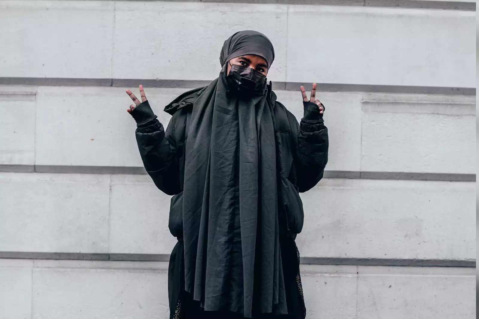 A young woman is wearing a grey hijab, puffer jacket and mask makes a peace sign with both her hands. She is stood in front of white stone bricks. (The young woman, Ebyan, appears in all of Saidul’s photographs here.)