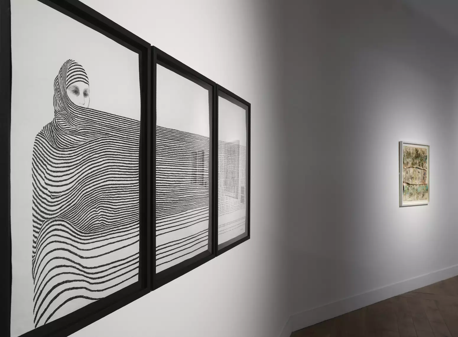 A gallery shot showing a striking black and white drawing of a person wrapped in a flowing striped headscarf and full-body garment, which extends across three black frames on a white gallery wall. The persons eyes and nose show above the fabric; their eyes are shaded as if sheltered from the sun, or because they are wearing smoky eye shadow.