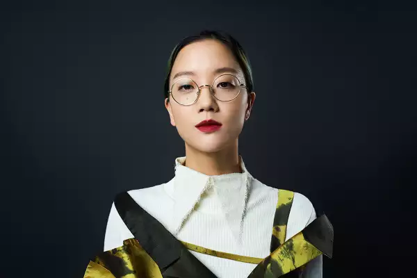 A head and shoulders portrait of the artist Christine Sun Kim. She wears her hair pulled back, round glasses and burgundy red lipstick with an off-white top with a pointy collar and a gold and black dress against a plain, dark background. She is looking directly into the lens.