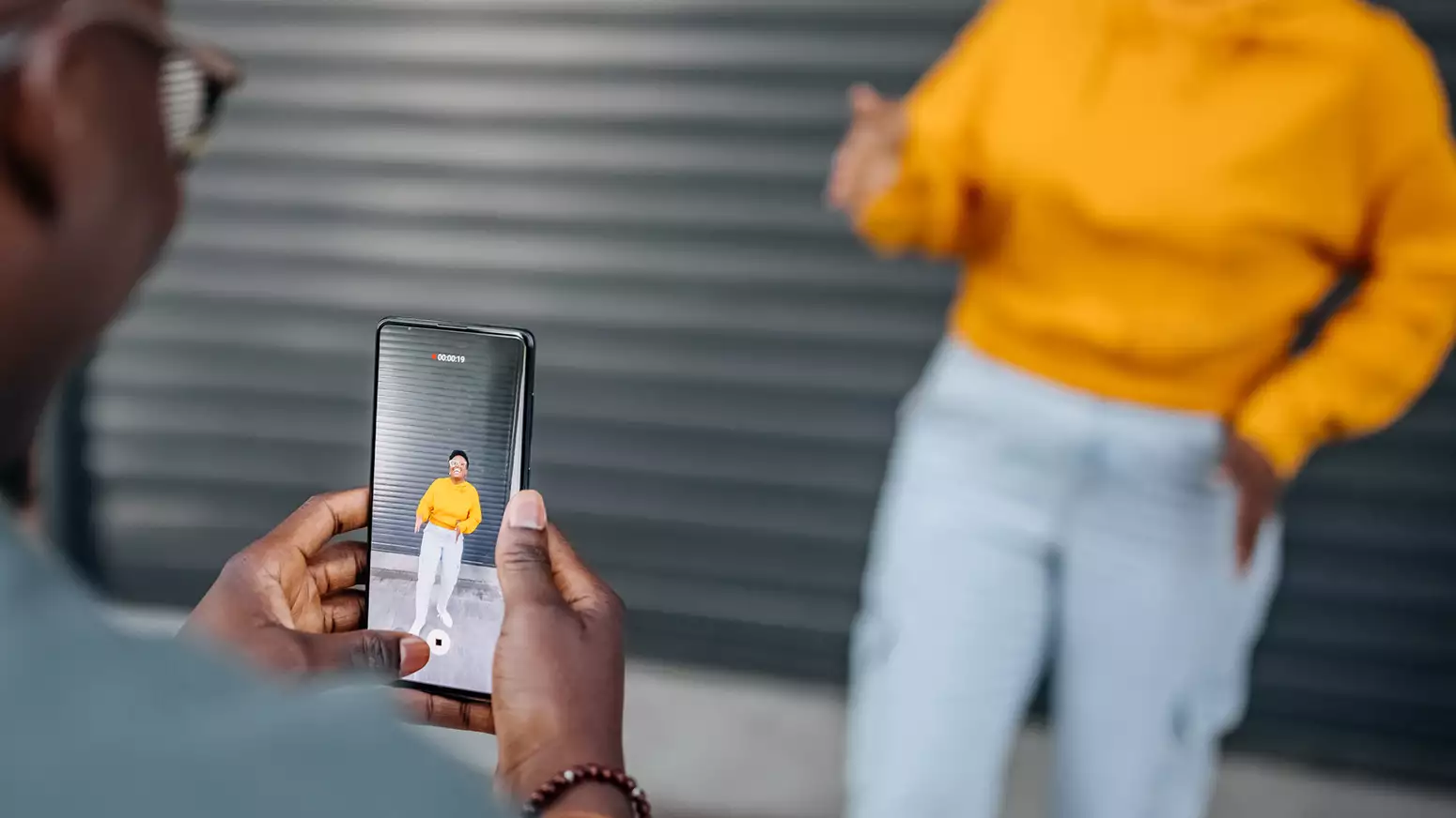 A person posing against a metal shutter looking happy, whilst someone takes their picture on a mobile phone