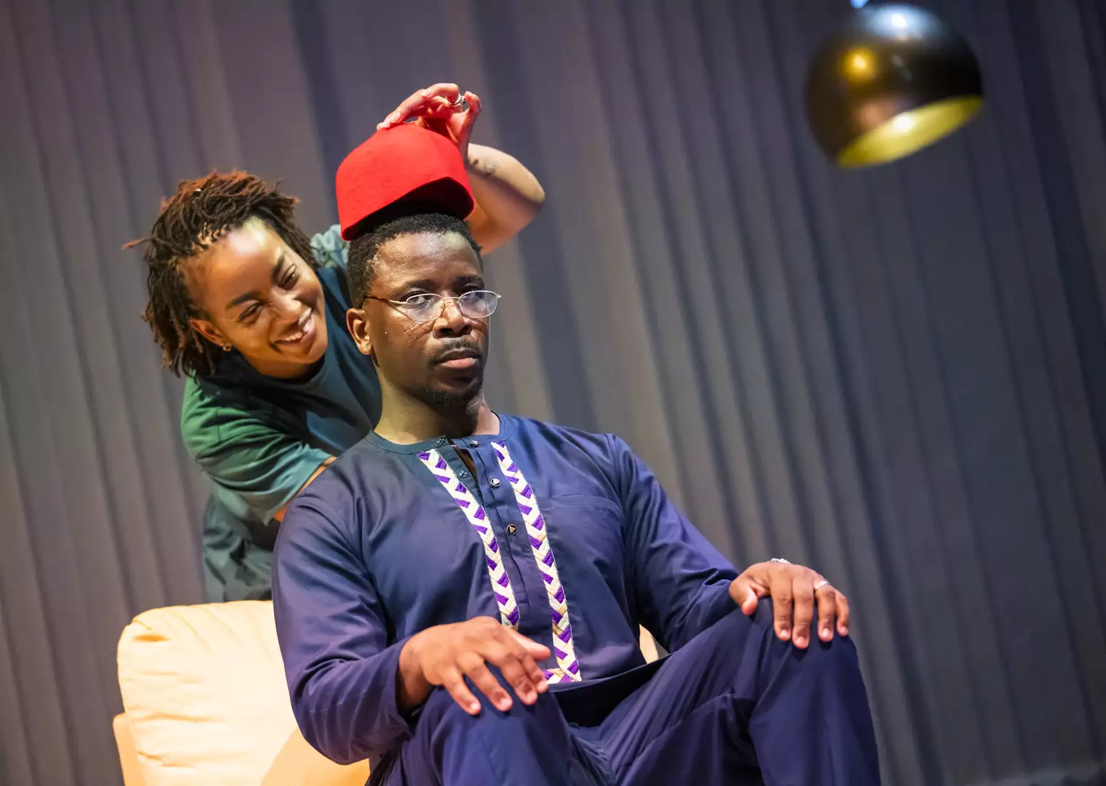 Uche Abuah and Itoya Osagiede on stage in Notes on Grief. Osagiede is Black with dark brown skin, short black hair, moustache and goatee beard; he sits on a yellow armchair wearing a blue dashiki with white and purple details over blue trousers, he wears glasses and has a serious expression. Abuah stands behind him and laughs as she holds a red hat above his head. Abuah is Black with medium brown skin; her hair is in fine locks, pulled back; she wears a blue T-shirt and trousers.
