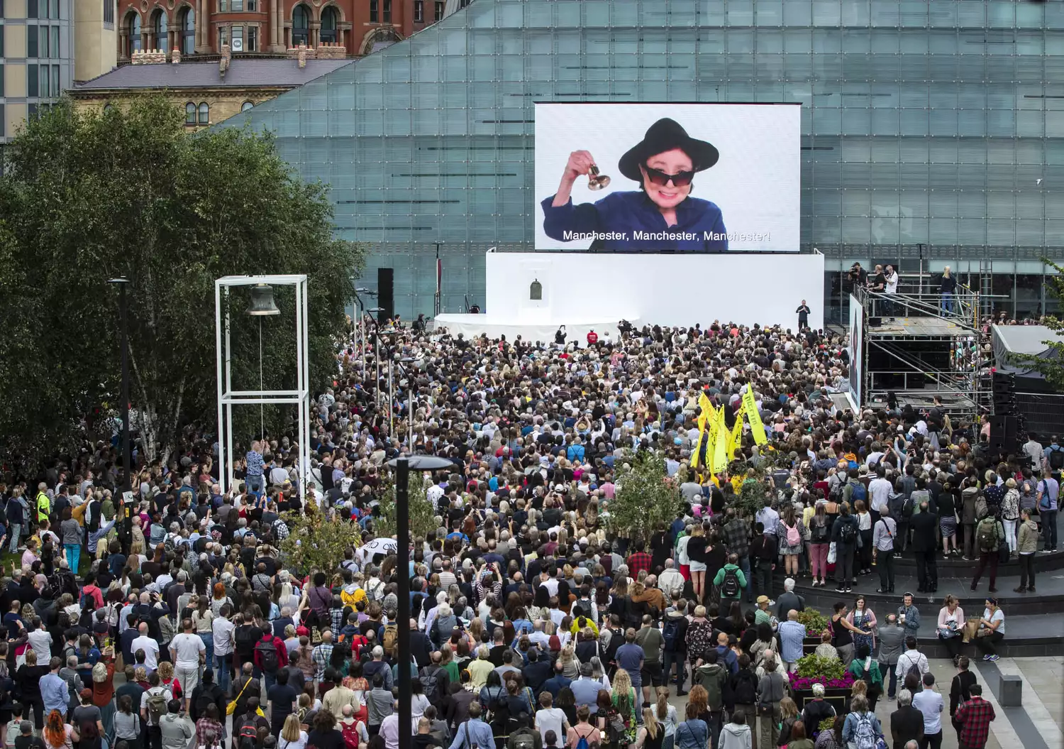 A large crowd of people are gathered holding hand bells. A screen in front of the crowd shows the artist Yoko Ono.