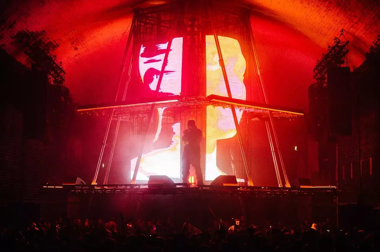 A photograph of Skepta, stood in silhouette on a brightly lit stage. The stage is a custom-built pyramid with a flat top, with a centrepiece screen showing 3D graphics of a human head shown across multiple large LED displays