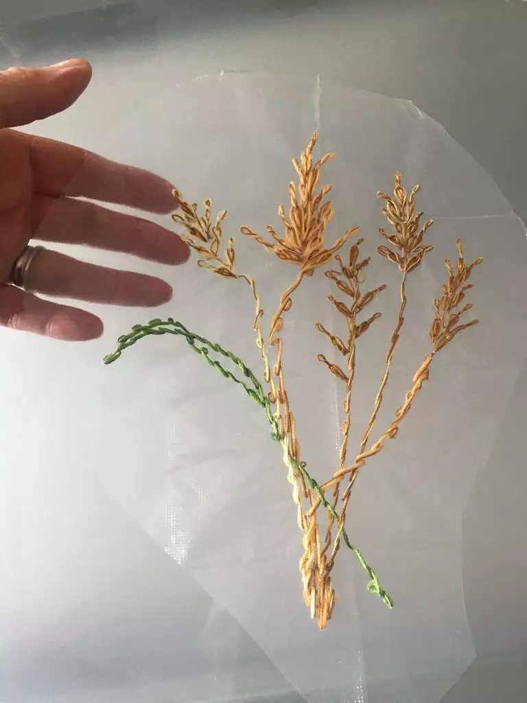 Gold and green grasses embroidered onto a sheer backing fabric, held up by a hand with a pale peach skintone.