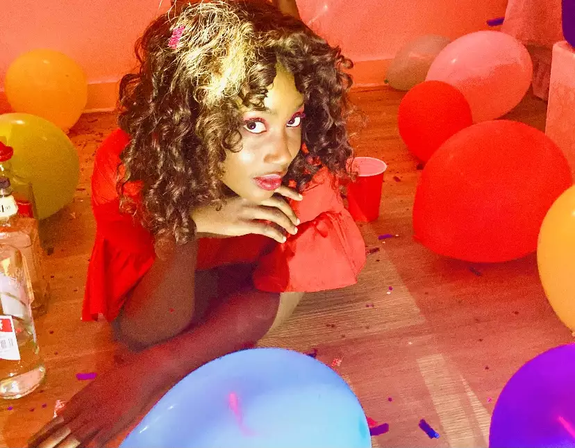 Bitez is looking up at the camera, resting her head on her hand. She has brown curly hair which is flecked with sunlight, she is wearing a red top, has red lipstick on and pink eyeshadow. She is surrounded by balloons, the floor is covered in confetti.