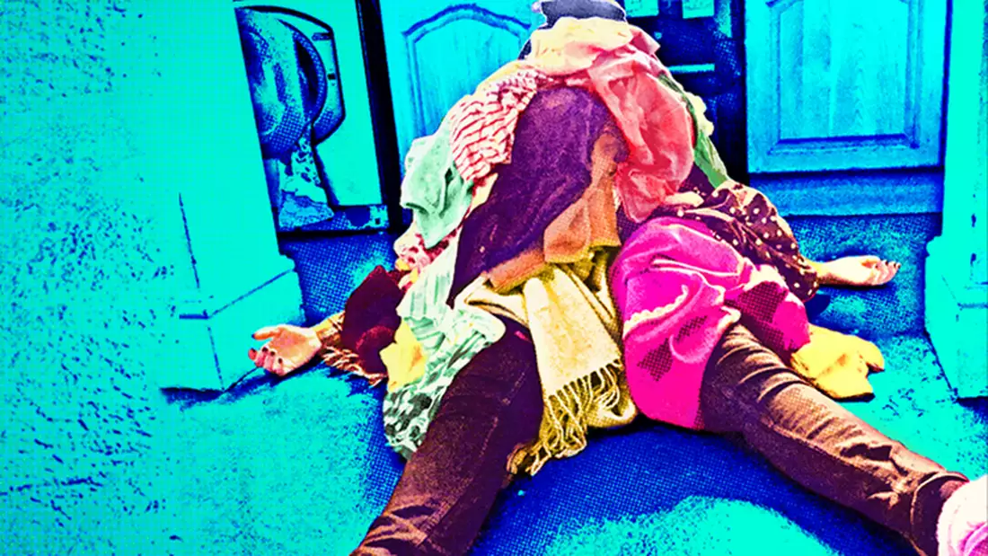 Woman lying on the floor of a kitchen with a pile oflaundry on top of her