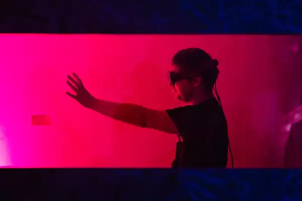 A man holds out his left arm in front of him, looking at his hand while wearing a Magic Leap mixed reality headset. The photograph is taken through a window, with the man standing in a small room lit with a bright pink wash