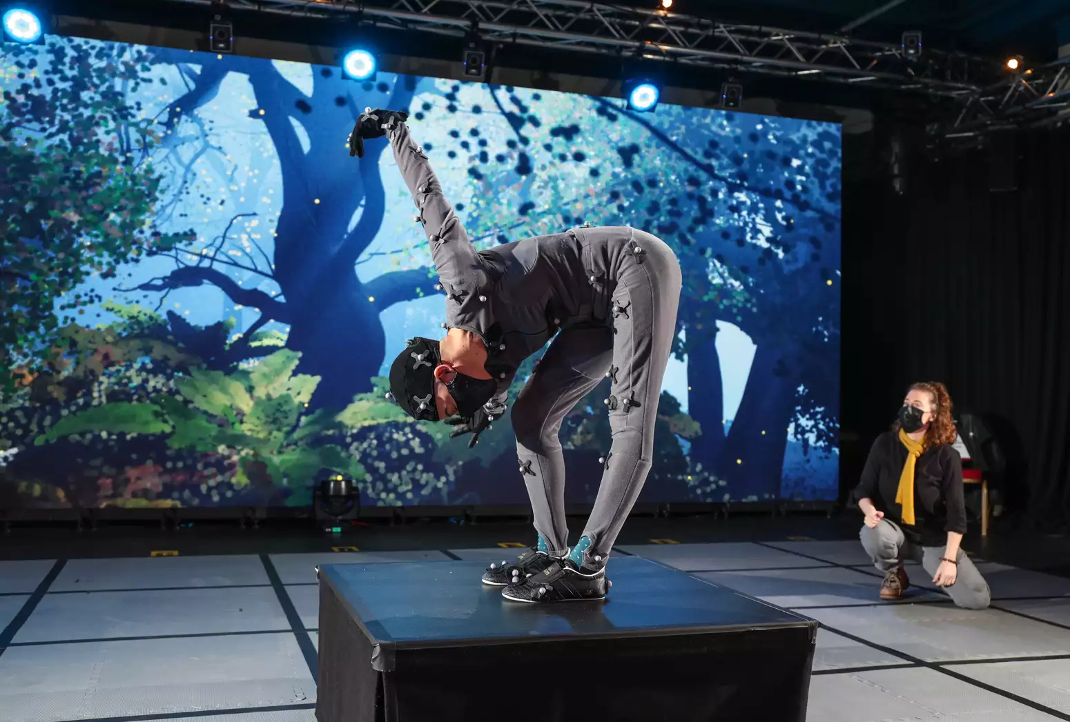 A person wearing a grey motion capture suit stands bent over at the waist in front of a digital screen