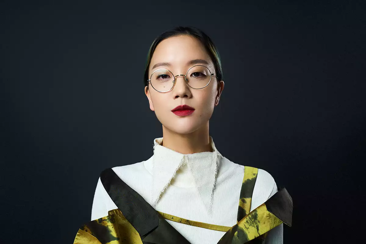 A head and shoulders portrait of the artist Christine Sun Kim. She wears her hair pulled back, round glasses and burgundy red lipstick with an off-white top with a pointy collar and a gold and black dress against a plain, dark background. She is looking directly into the lens.
