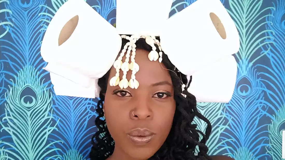 Artist Chanje Kunda standing in front of a blue background, wearing a headdress made out of toilet rolls