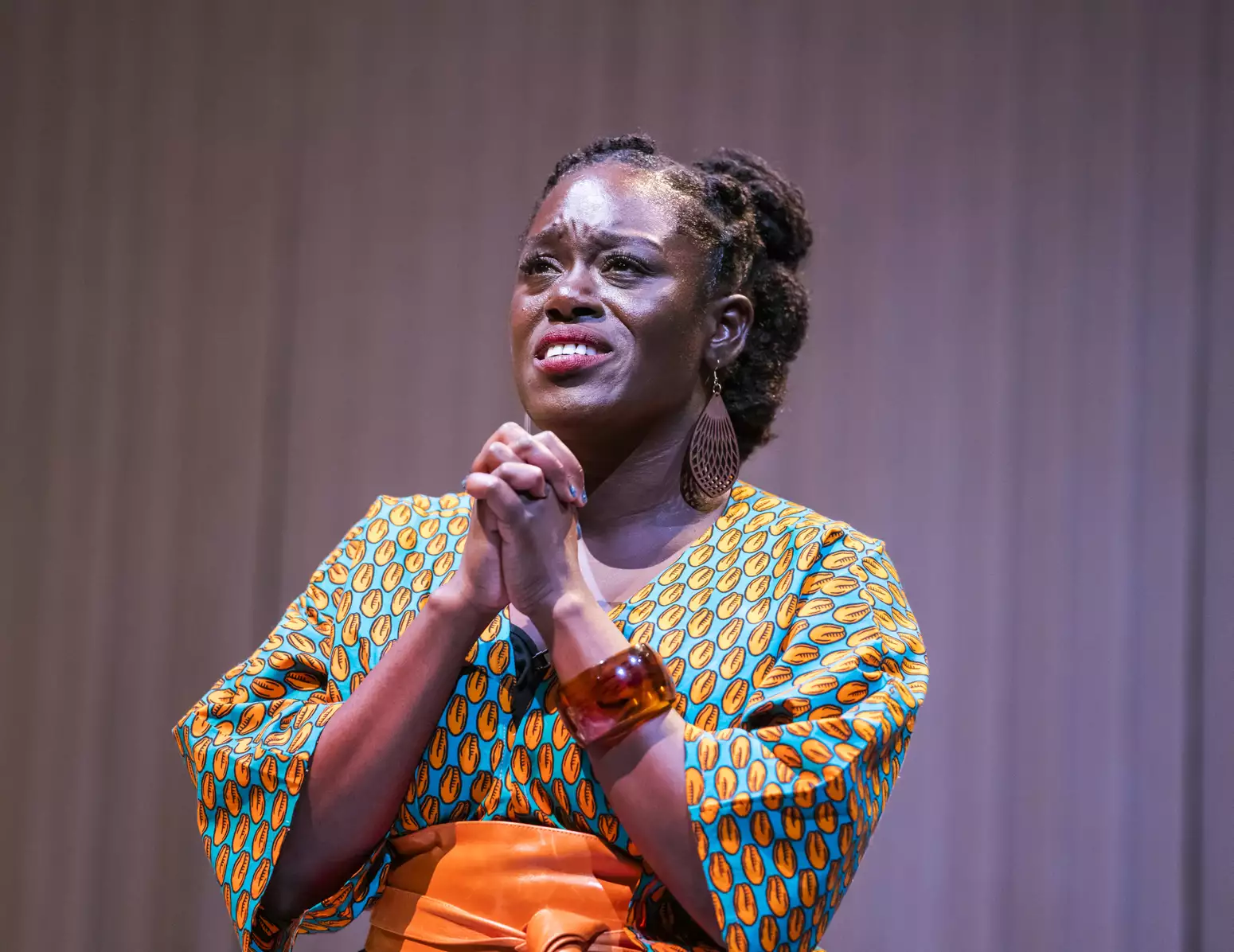 Michelle Asante on stage in Notes on Grief. Asante stands with her hands pressed together with fingers intertwined, her brow is furrowed as if in pain, her eyes look upward as if in prayer. Asante is Black with dark brown skin; her hair is twisted from the crown into three buns at the back of her head; she wears a bright orange and turquoise patterned dress and an orange wrap belt.
