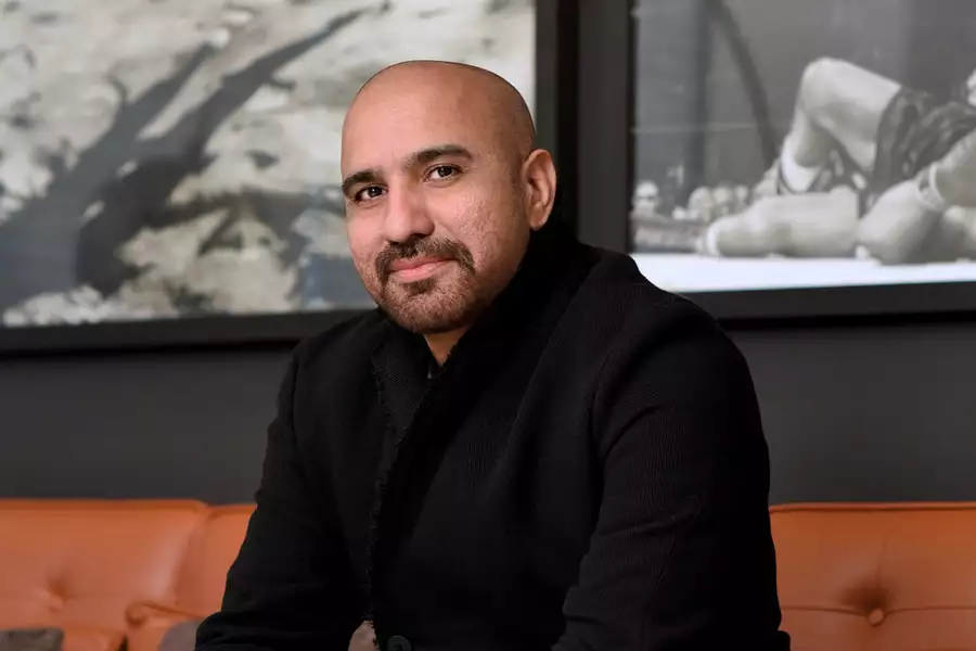 Photographic portrait of Rashid Rana sat on a sofa in front of 2 framed black-and-white photographs