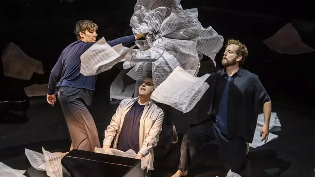 A photograph from Tao of Glass, showing Phelim McDermott sat at a miniature grand piano while two puppeteers construct an oversized wig for him using large pieces of sheet music.