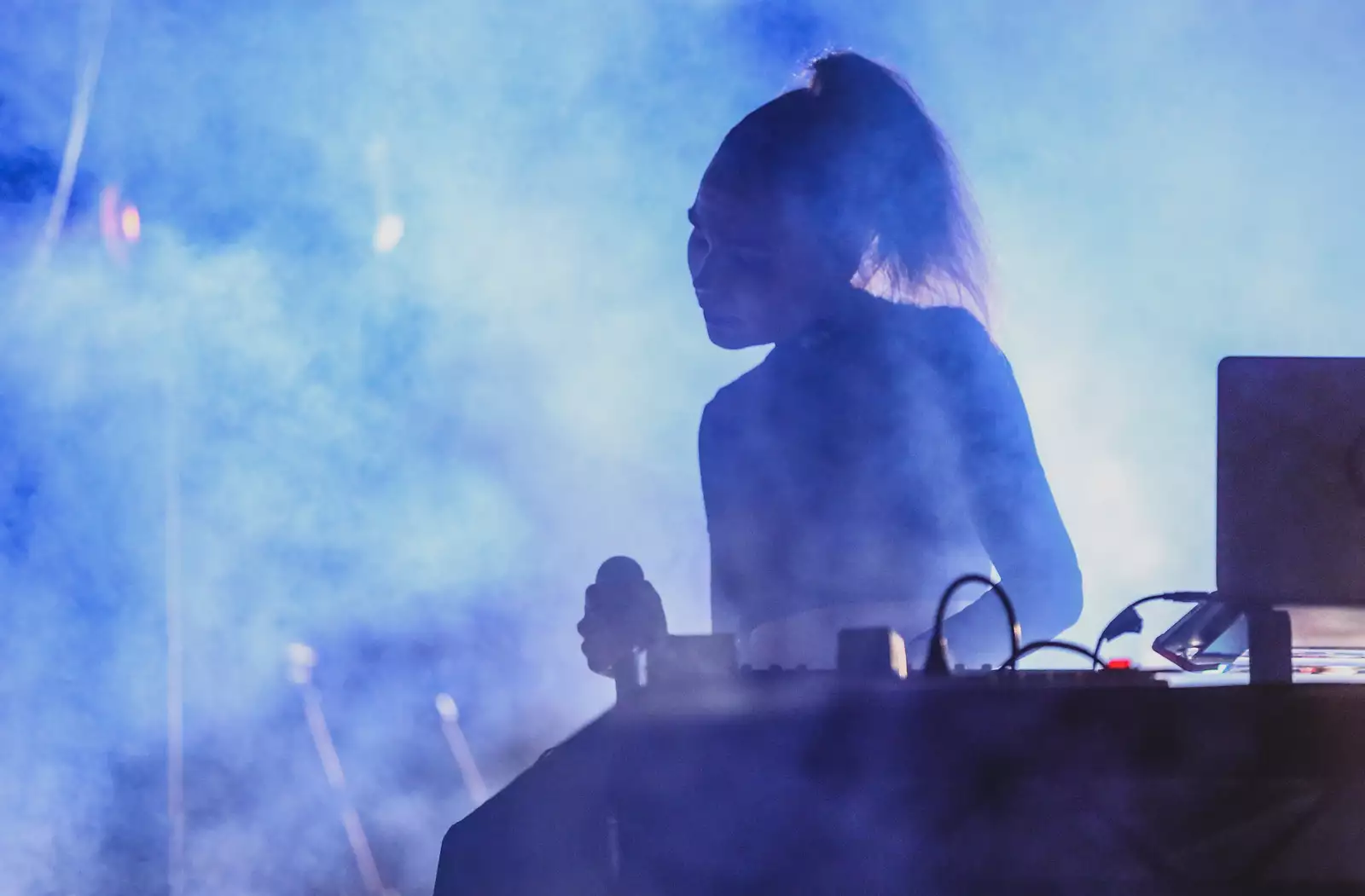 A woman onstage stood behind a laptop and controller holds a microphone, surrounded by dry ice