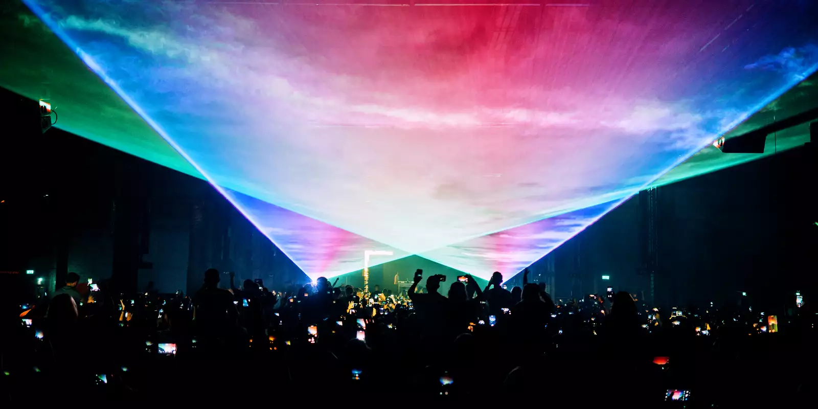A photograph of a large crowd at a concert, with many phone screens visible as they photograph the stage. The room is dark apart from two large, triangular fields of laser and smoke filling the top half of the image.