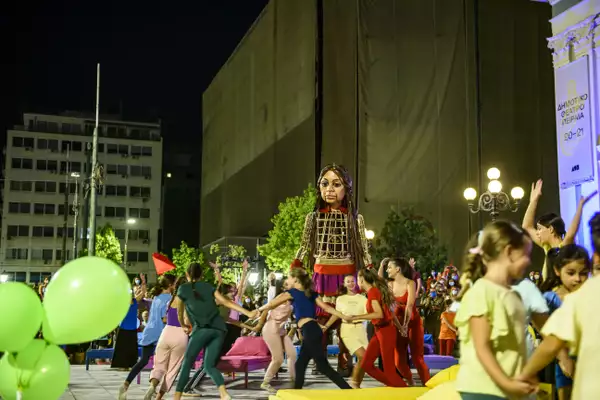 Groups of child performers run in rings holding hands while the Little Amal puppet looks on. It's night-time but the square is lit up brightly with street lamps, and is decorated by bright green balloons, colourful low beds and a crowd of people wearing face masks and taking photos on their phones.