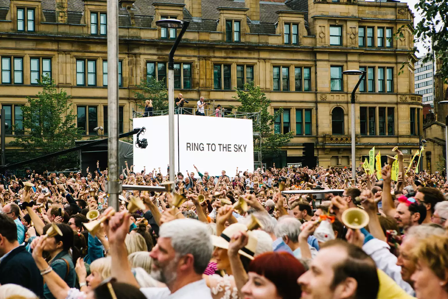 A crowd of people ring bells. In the middle of the crowd, there is a white banner with 'RING TO THE SKY' written in black text.