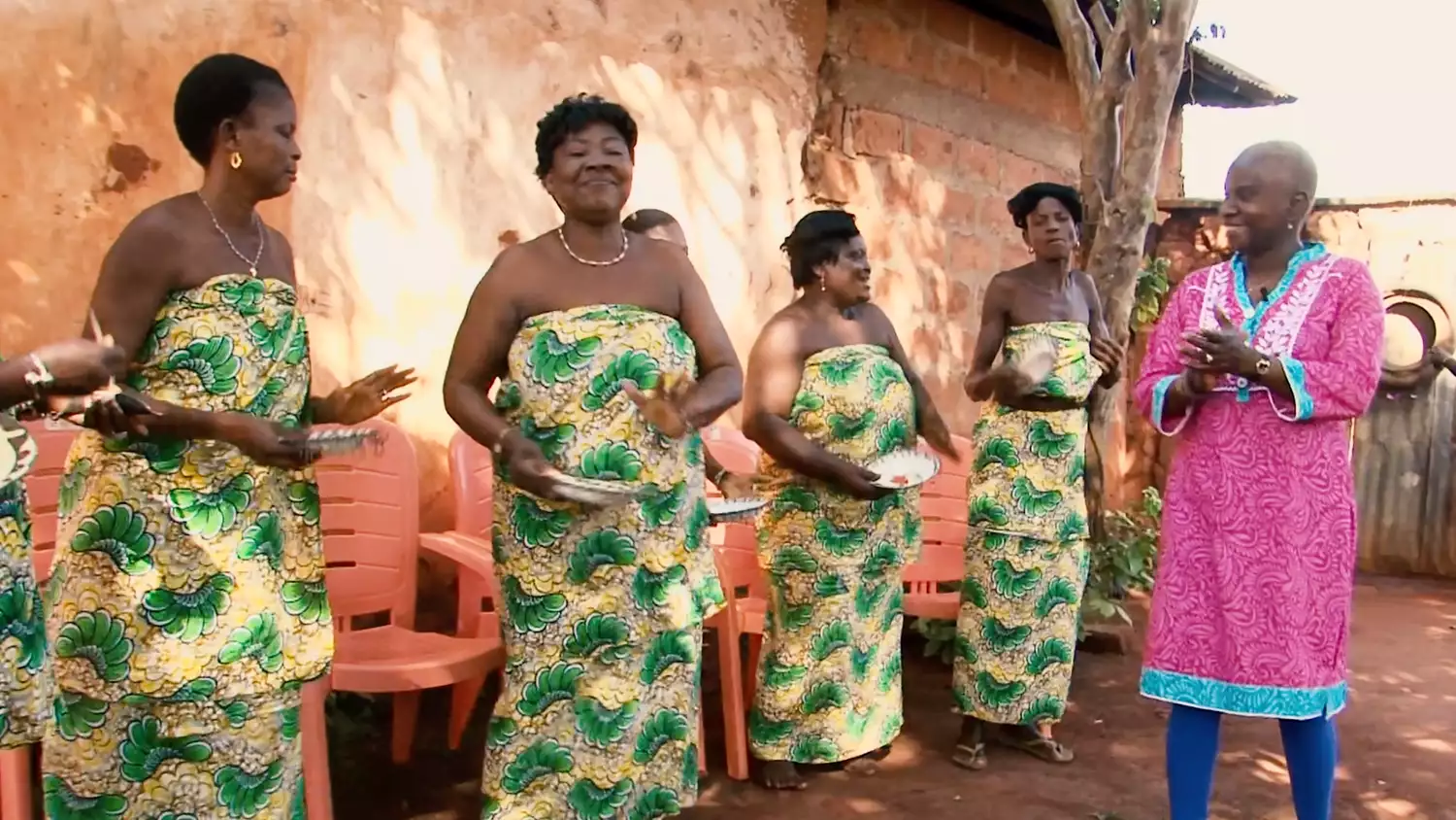 A photograph of four women in Benin, wearing matching pineapple print dresses and standing mid-song. A fifth woman wearing pink appears to conduct them.