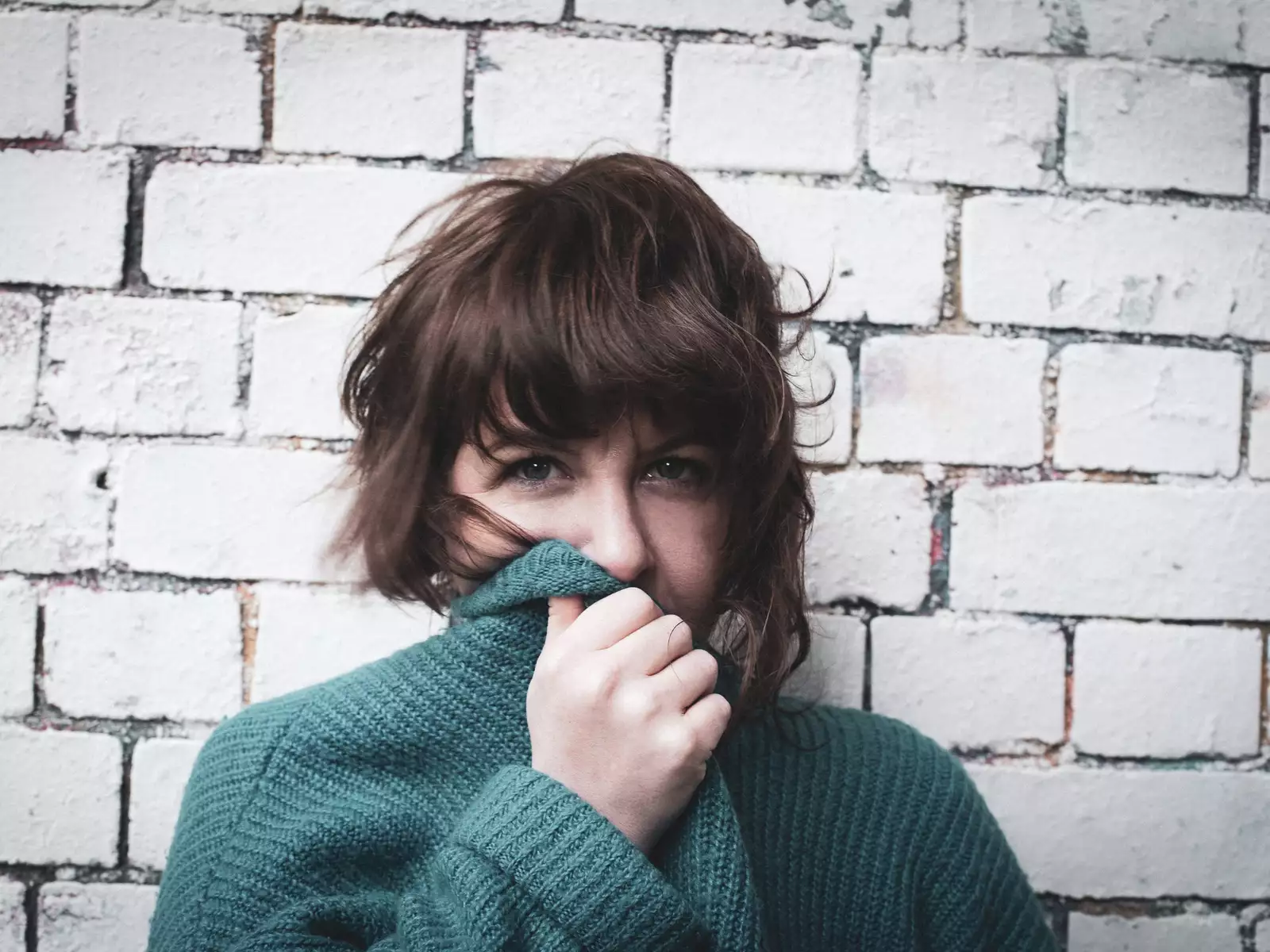 Facing directly into the camera  you can see that Test Card Girl is wearing a green cardigan or jumper, she is partially covering her face with the collar of it. She has wavy brown hair and is standing in front of a white brick wall.