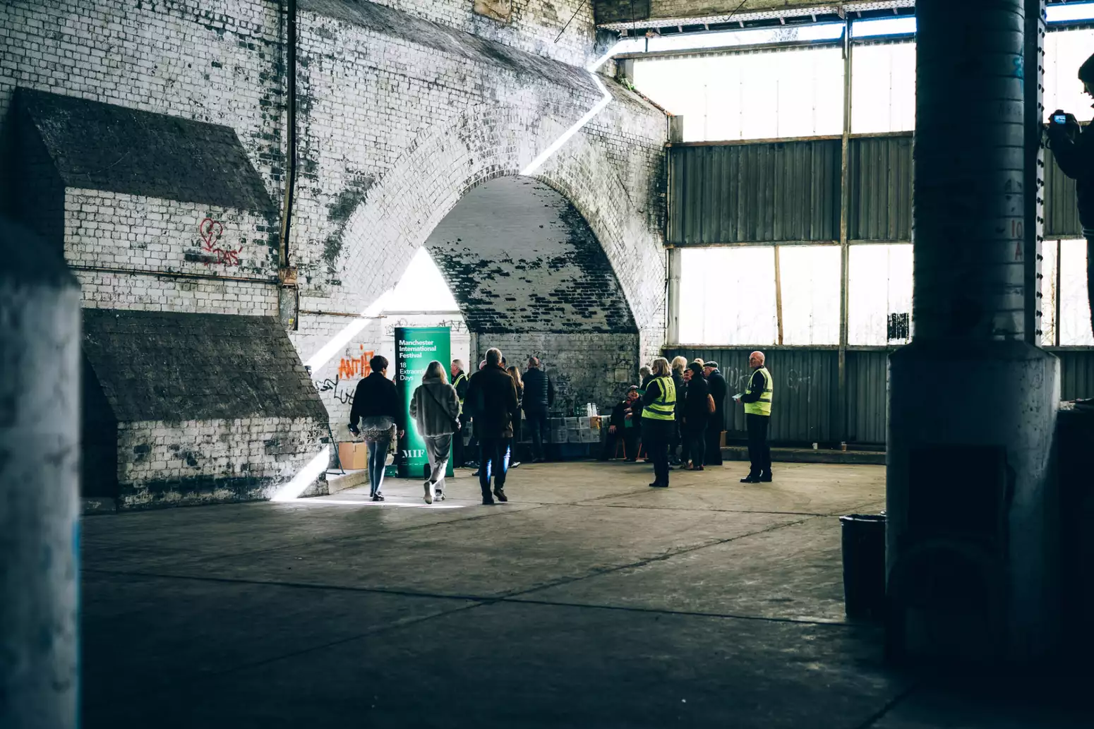 A small crowd of people are gathered in a large room with a brick facade on one side and windows on the other.