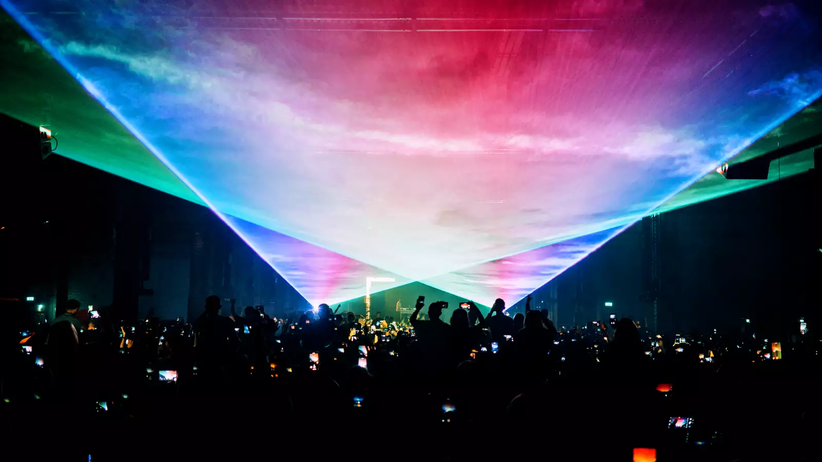 A photograph of a large crowd at a concert, with many phone screens visible as they photograph the stage. The room is dark apart from two large, triangular fields of laser and smoke filling the top half of the image.