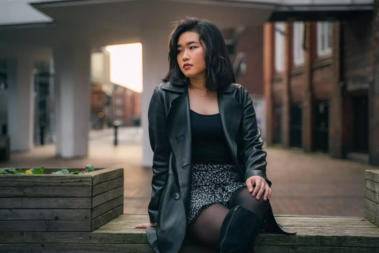 A woman is sitting on a wooden bench, body facing towards the camera but she is looking off to the left into the distance. She is wearing a long black jacket, black top and flowery skirt.