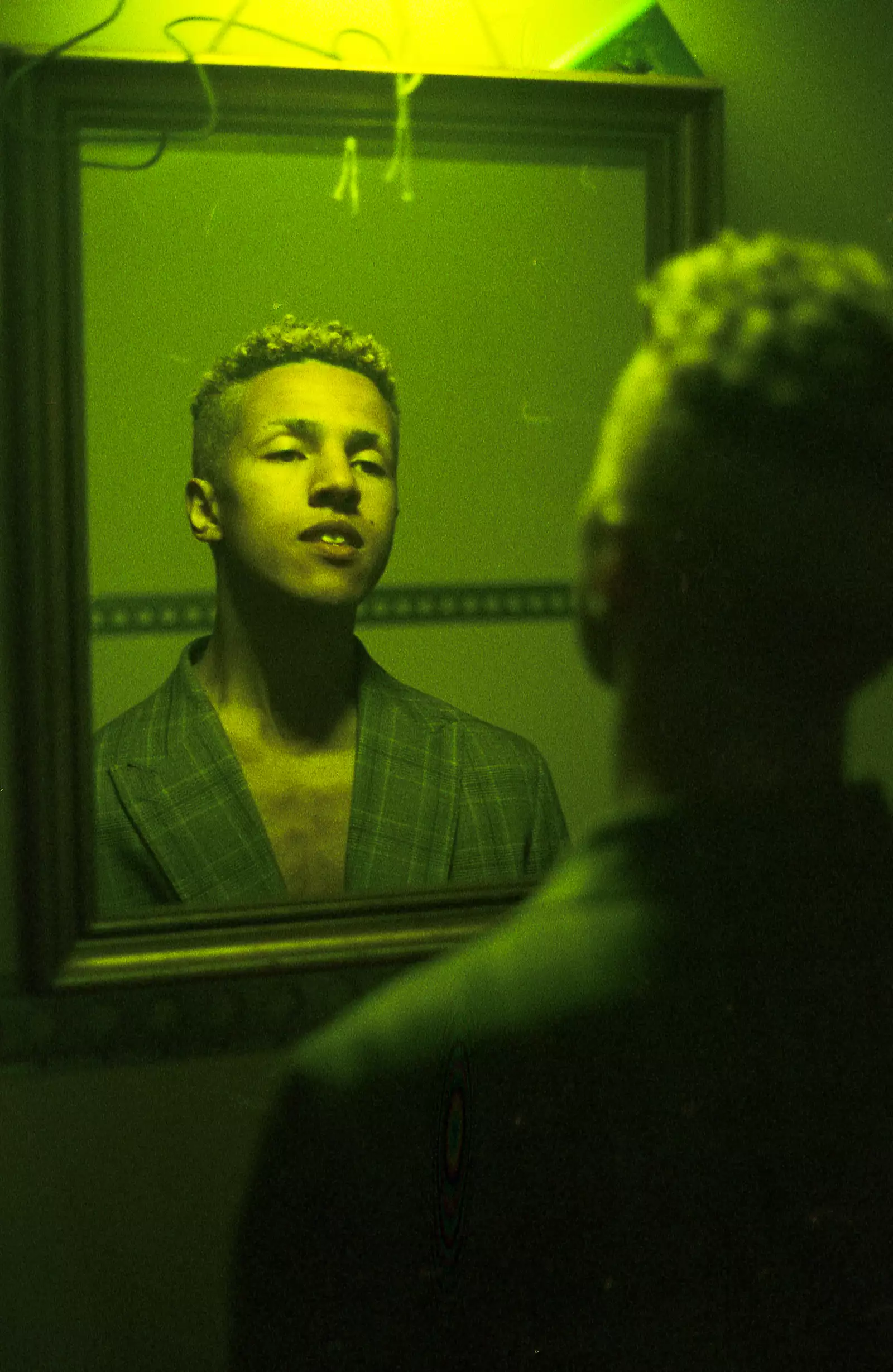SIBZ is looking into a mirror. They have bleach blonde tight curls and shaved sides. They are wearing a suit blazer, the image has a green tint to it.