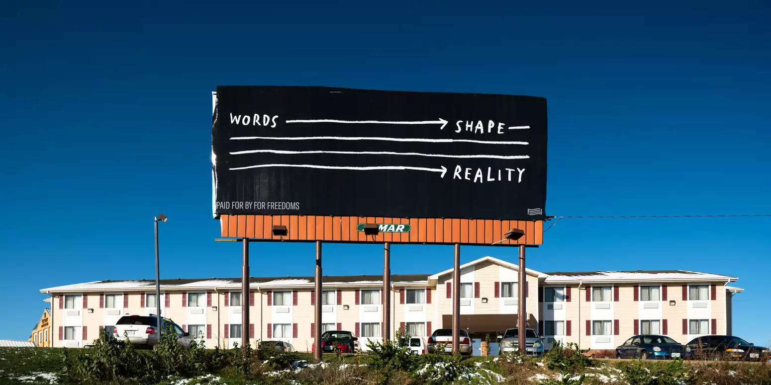 An image of a billboard high in the blue sky over a motel. It has a black background and white text that reads "WORDS" then a white arrow to the text "SHAPE" then a white arrow that runs over three lines to the text "REALITY"