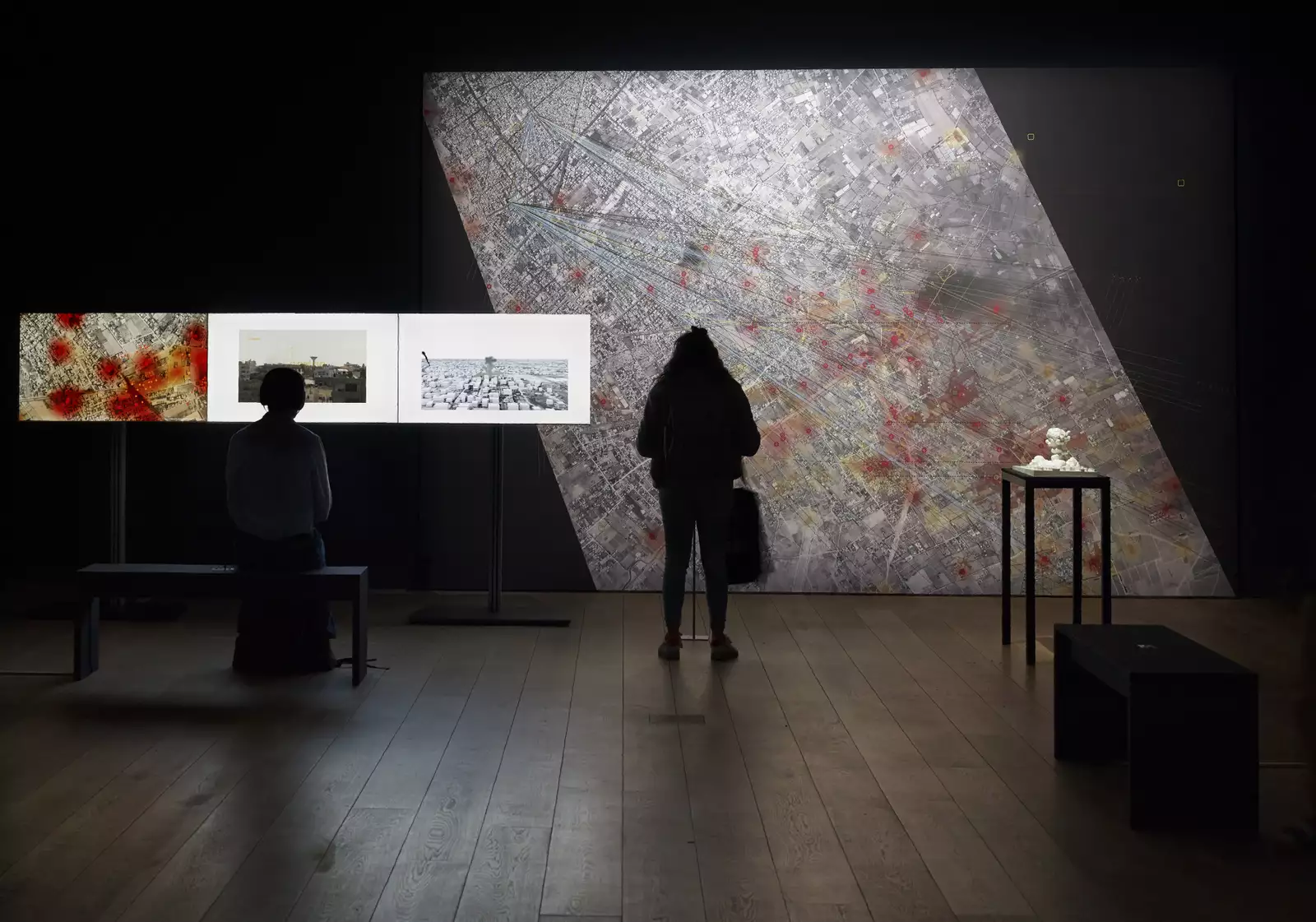 A photograph of two people stood in silhouette inside an art gallery - one is looking at a large map covering a wall, while the other watches a multichannel video installation on 3 screens.