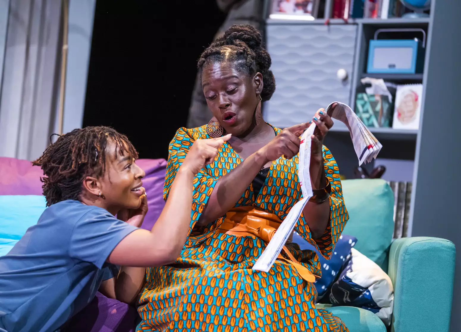 Uche Abuah and Michelle Asante on stage in Notes on Grief. Asante sits on a blue and purple sofa and holds up a folded paper map, which she points to. Abuah sits at her feet, smiling and also points at the map. Asante is Black with dark brown skin; her hair is twisted from the crown into three buns at the back of her head; she wears a bright orange and turquoise patterned dress and an orange wrap belt.  Abuah is Black with medium brown skin; her hair is in fine locks, pulled back; she wears a blue T-shirt.