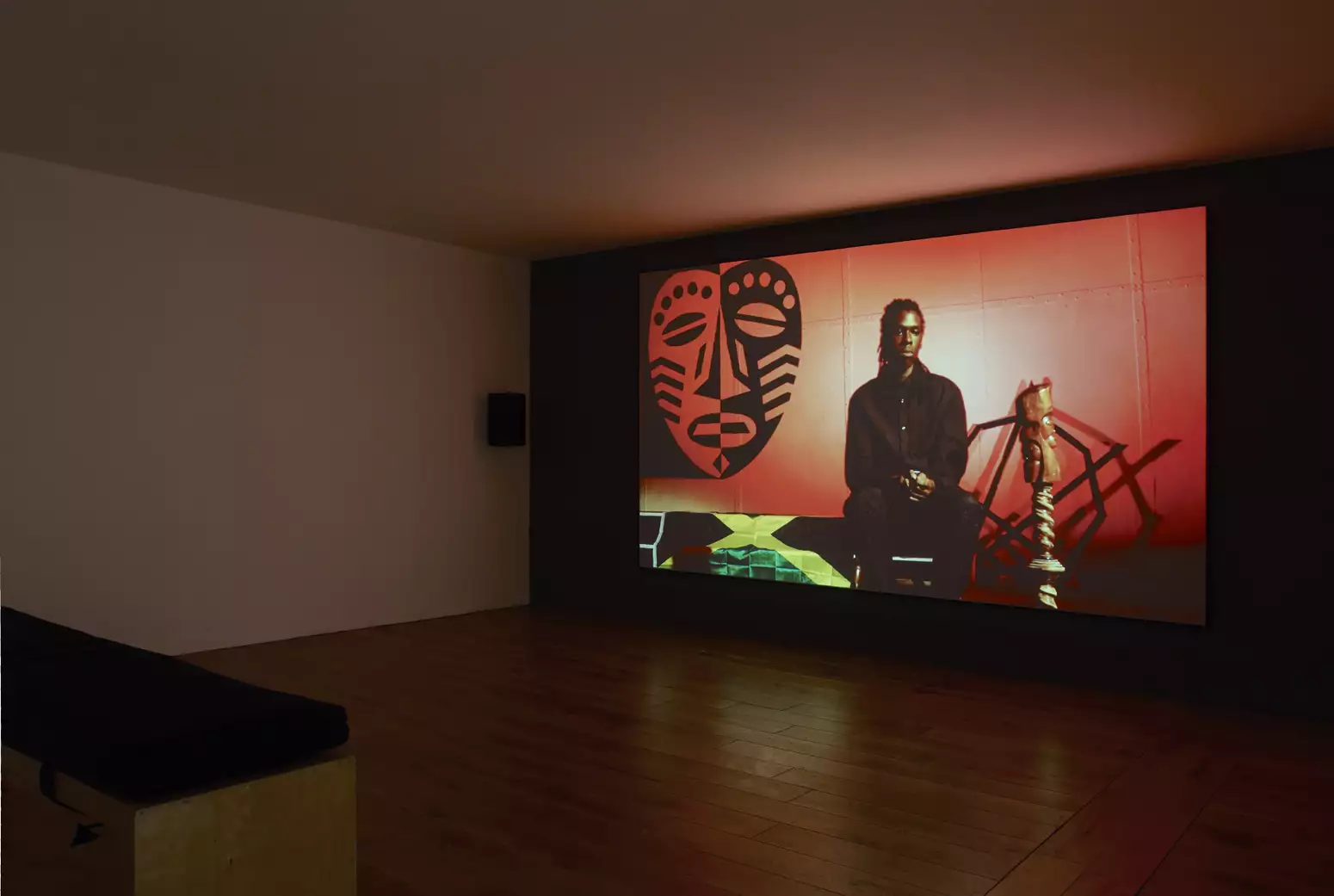 An image is projected onto the back wall of a gallery space. The image has a red background; a Black person with a dark brown skin tone and braided hair is seated in the centre; they are wearing a black button-up shirt and black trousers; they hold their hands together in their lap; light shines on their skin, casting it in a gold hue; they have a thoughtful expression. In the top left corner of the image is a very large mask in black and red; each side is a reverse mirror image of the other; the mask has dots for eyebrows, coffee-bean shaped eyes, stripes on its cheeks and marks beside its nose and on its chin. It has a straight mouth shape. Under the mask, the flag of Jamaica is laid out. On the right side of the image is a wooden sculpture with a twisted stem holding up a what looks like another form of mask.