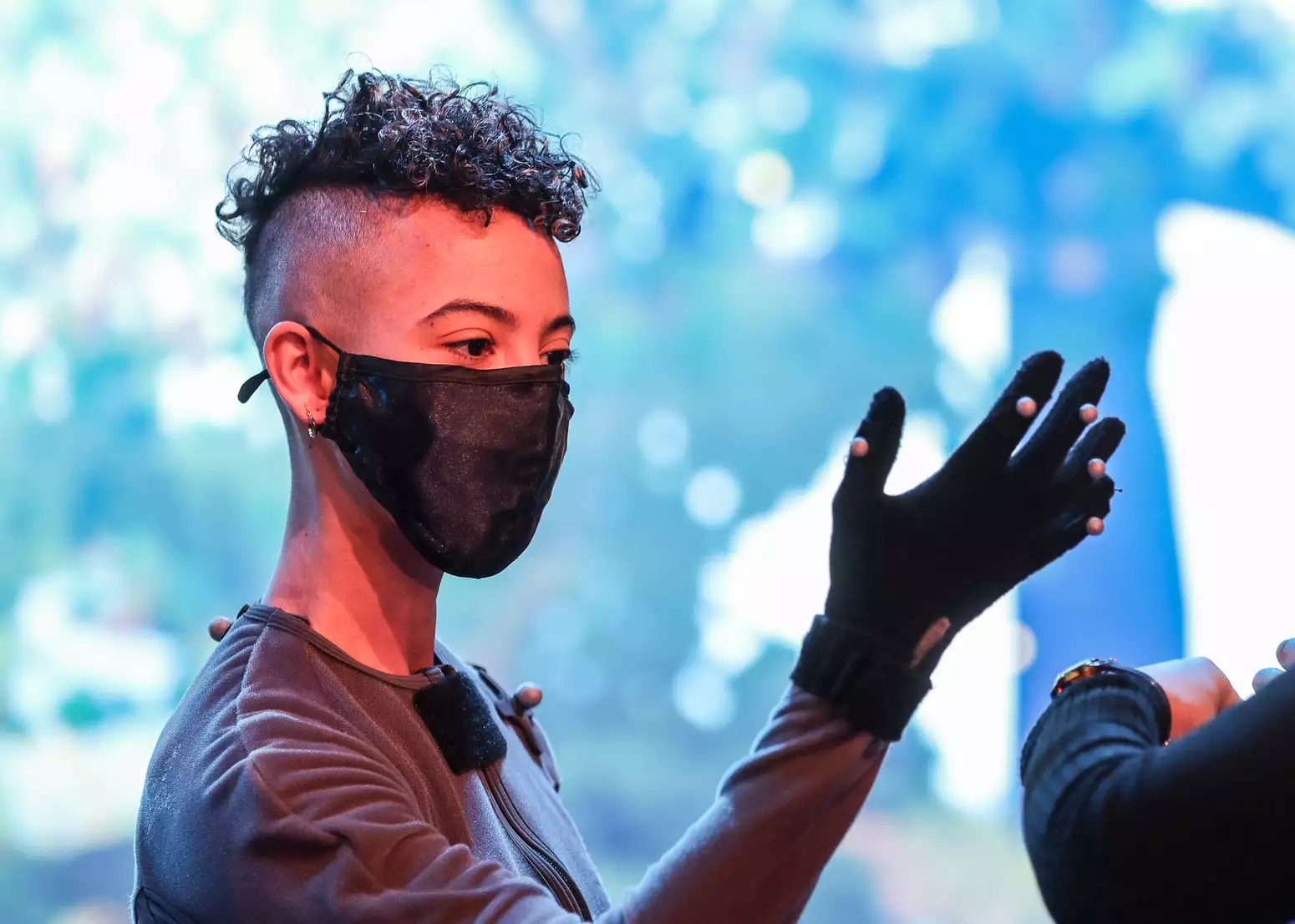 A person with curly black short hair with shaved sides wears a black mask and a grey motion capture suit while standing in front of a digital screen