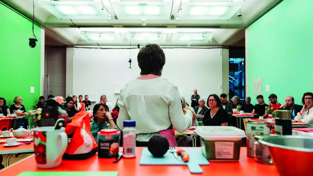 A photograph of a room at Manchester Art Gallery which has been converted into a temporary classroom, with bright green walls on the left and right of the frame. A class of adults are listening intently as the tutor, with her back to us, leads a cookery class.