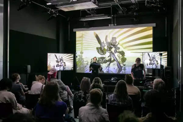 A workshop of sitting participants, a man with blonde hair is delivering a workshop in front of a digital screen