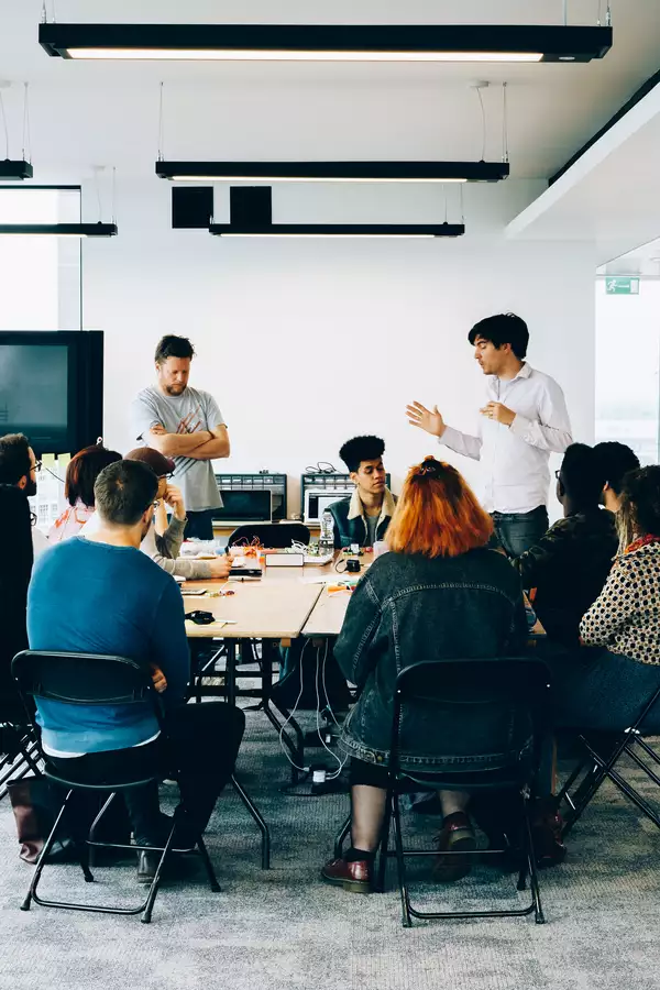 A number of people are sat around a large table while two people are stood speaking.