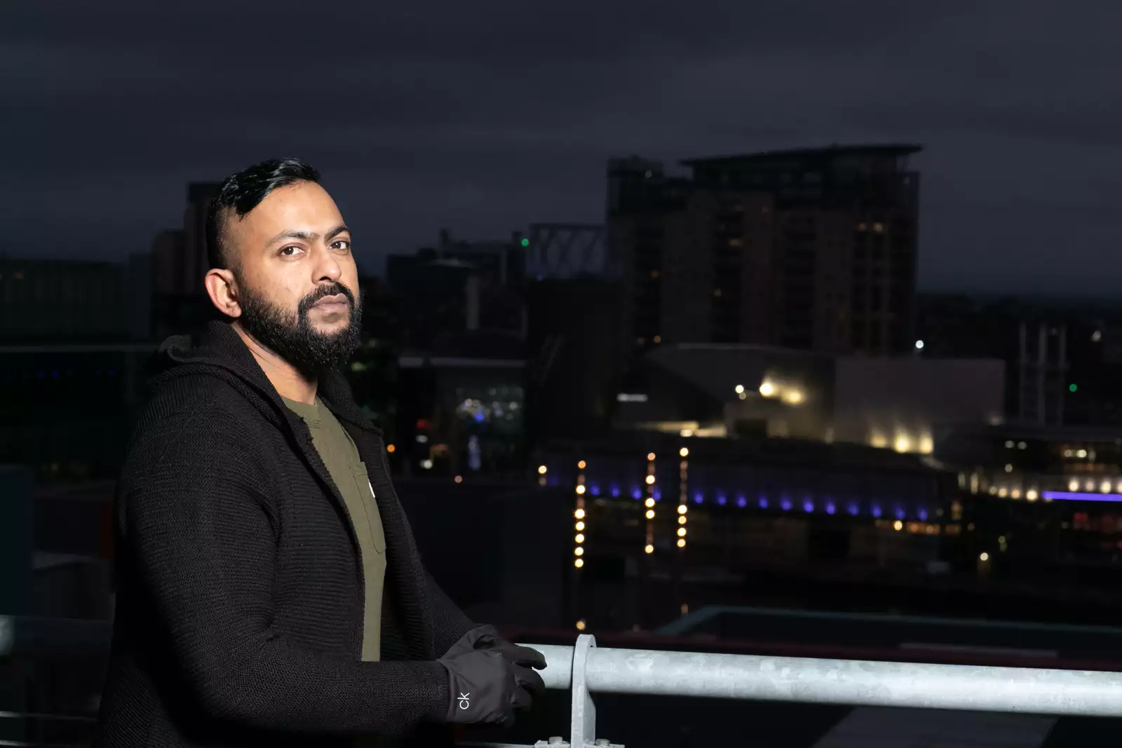 It's nighttime. Sam is stood on a bridge, side on, but looking at the camera. He's wearing a black jacket and gloves. There is a cityscape in the background