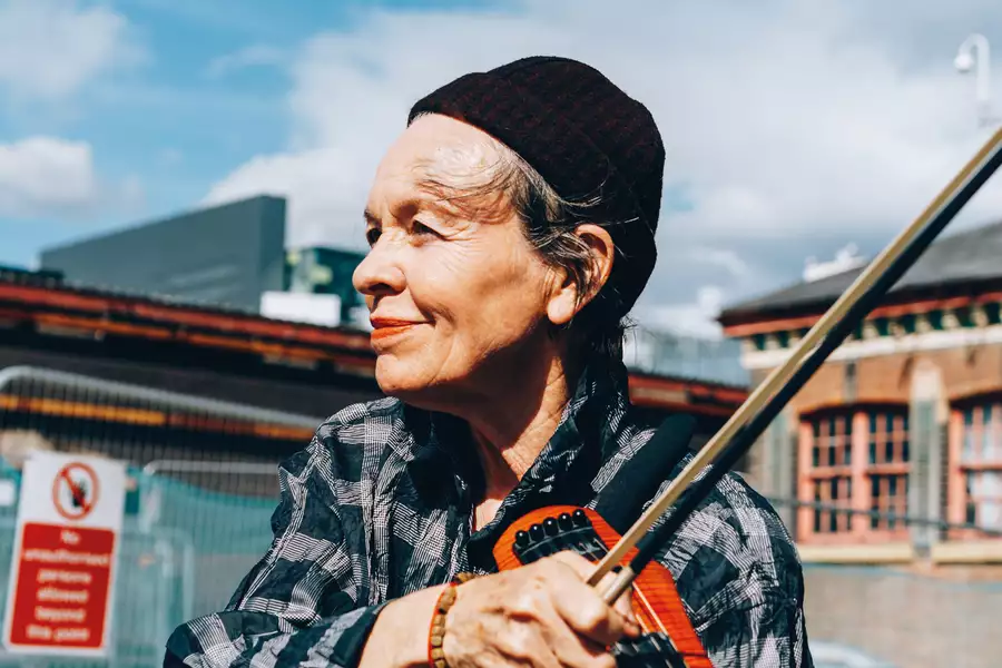 A photograph of Laurie Anderson stood outside at a construction site, plucking a string of an electric violin which is resting on her left shoulder,