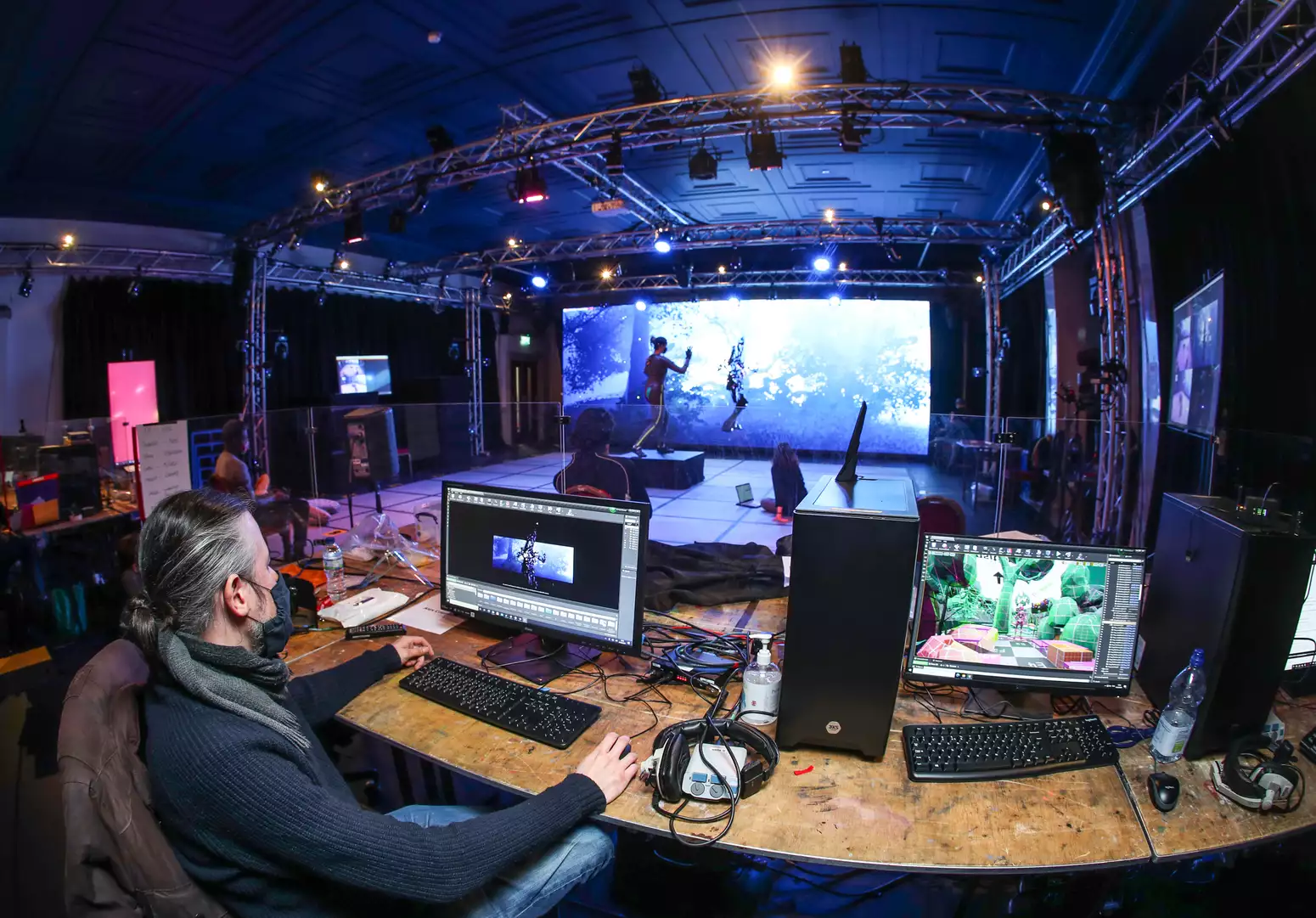 A virtual production studio is seen from behind a bank of computer screens, an actor in a motion capture suit is seen on the stage, their posture mirrored by an avatar on a digital screen behind them