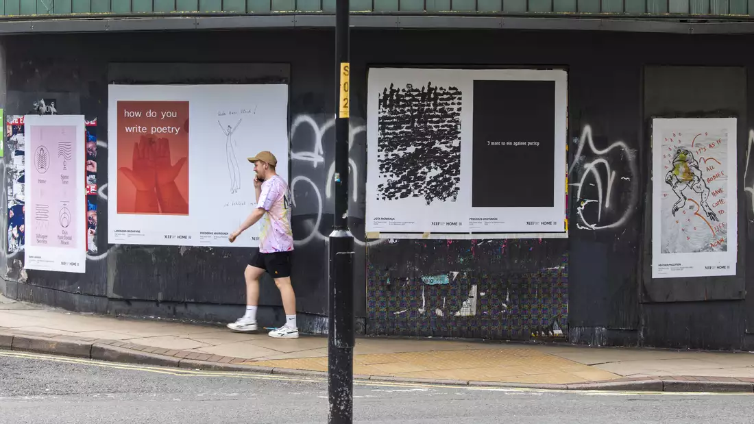 A photograph of a man walking past 4 posters showing artwork from Poet Slash Artist while talking on the phone.
